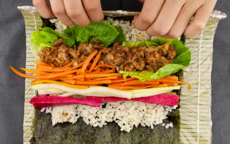 Trải tầm màn tre lên bàn, đặt lớp rong biển lên trên, dàn cơm rồi cho các nguyên liệu theo thứ tự bạn thích